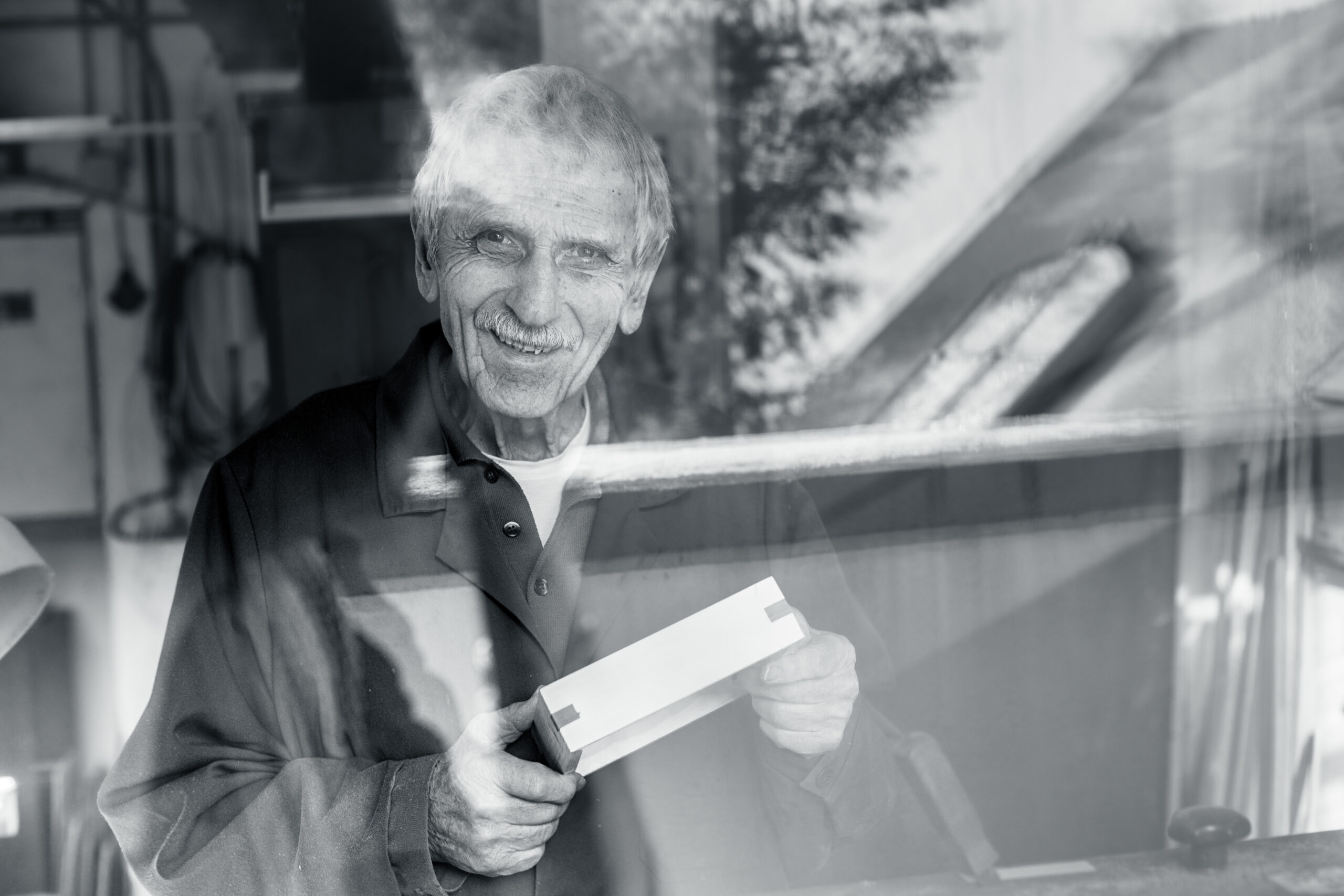 Projekt Forever Active - ein Tischler aus Leidenschaft, durch das Fenster fotografiert mit einem Holzstück in der Hand