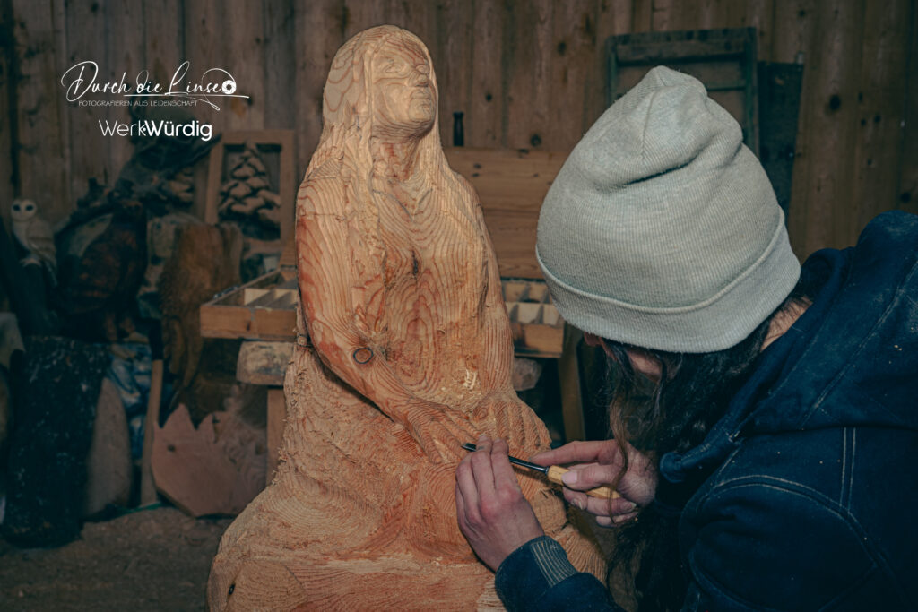 Holzkünstlerin Lisa Hirschbichler arbeitet mit einem Werkzeug an den Händen einer Holzskulptur.
